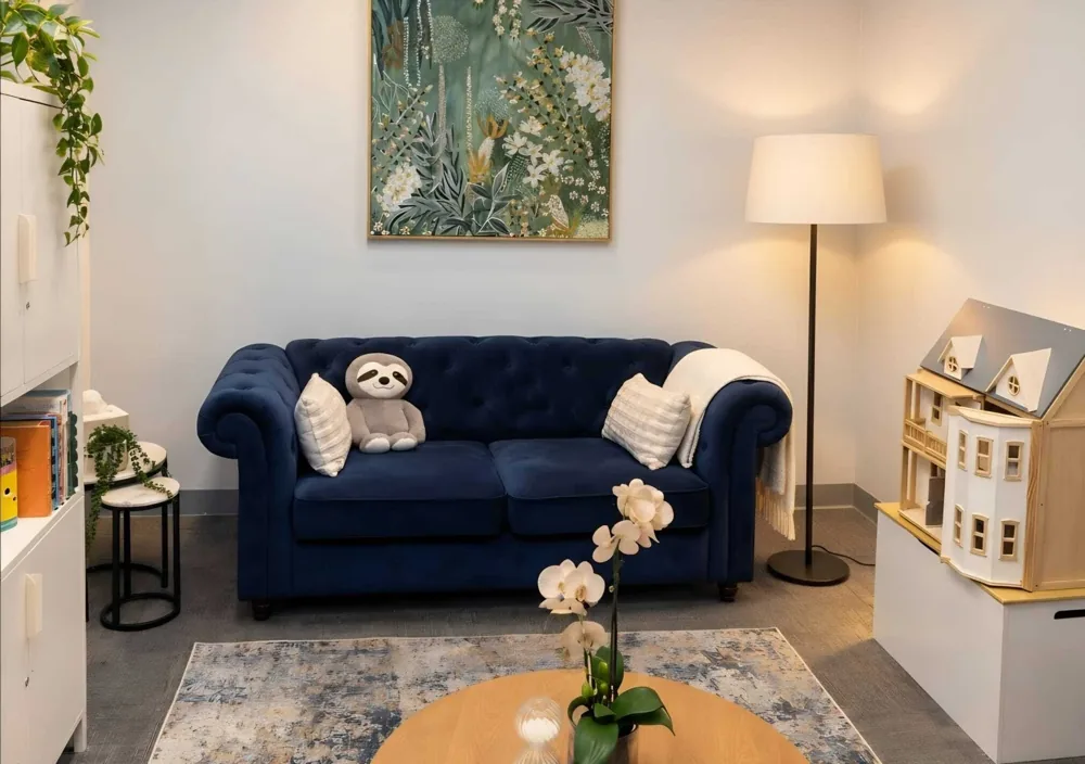 Calm consulting room with a navy sofa, soft lamp light, plants and a toy dolls house