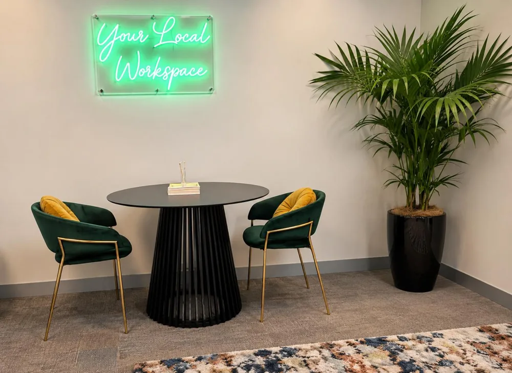 Two green velvet chairs and a round table beneath a neon sign, beside a tall palm